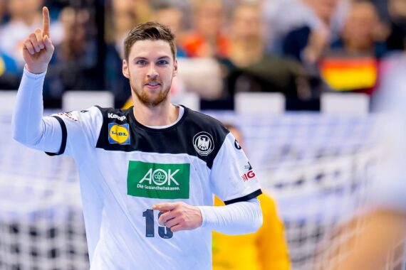 Ein männlicher Handballspieler, der ein weiß-schwarzes Trikot mit der Nummer 18 trägt, hebt auf dem Spielfeld den rechten Zeigefinger, während eine Menschenmenge und deutsche Fahnen im Hintergrund verschwimmen.