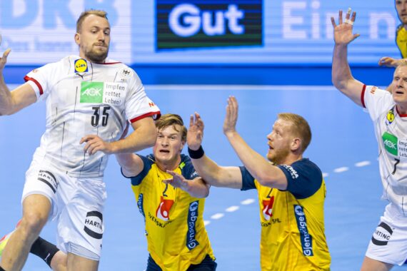 Ein Handballspieler in einem weißen Trikot springt, um den Ball zu werfen, während drei Verteidiger in Gelb und Blau versuchen, ihn zu blockieren. Im Hintergrund sind andere Spieler und ein blaues Spielfeld zu sehen.