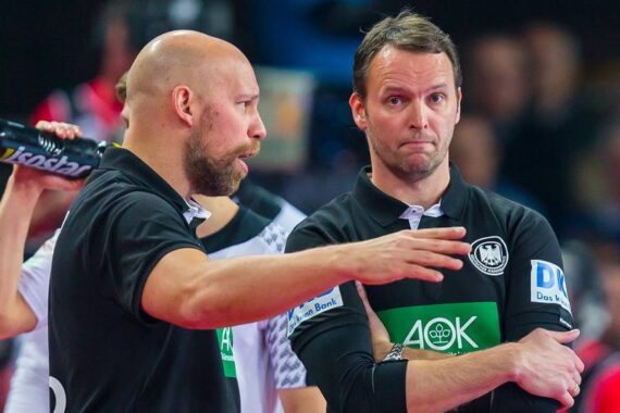 Zwei Trainer in schwarzen Hemden haben eine ernste Diskussion während einer Sportveranstaltung. Der eine gestikuliert mit seiner Hand und einer Wasserflasche, während der andere mit verschränkten Armen dasteht und unsicher schaut. Beide tragen Mannschaftslogos und Sponsorenaufnäher.