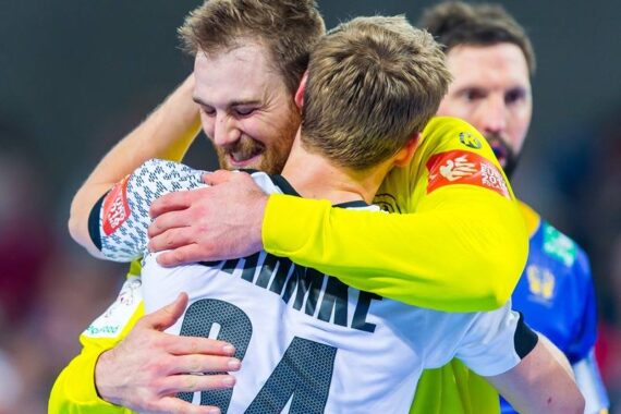 Zwei Sportler in Sporttrikots umarmen sich jubelnd, einer trägt ein gelbes Torwarttrikot, der andere ein weiß-schwarzes Trikot. Ein dritter Spieler in Blau steht im Hintergrund, leicht unscharf.