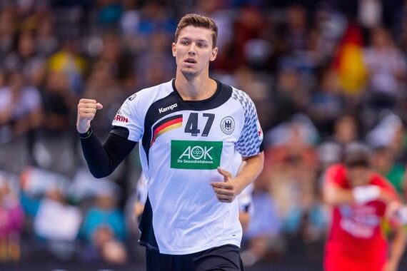 Ein männlicher Handballspieler, der ein weißes Trikot mit der Nummer 47 und einer deutschen Flagge trägt, macht eine Faust beim Jubeln während eines Spiels, mit einer unscharfen Menge und anderen Spielern im Hintergrund.