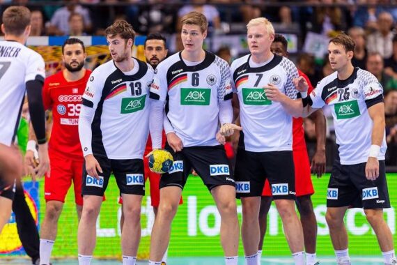 Vier männliche Handballspieler in weißen und schwarzen Uniformen stehen in einer Abwehrmauer und bereiten sich darauf vor, einen Freiwurf zu blockieren. Ein Spieler in Rot steht hinter ihnen und hält einen gelb-blauen Handball. Ein Publikum schaut aus dem Hintergrund zu.