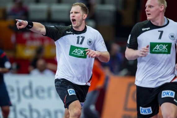 Zwei männliche Handballspieler in weißen Trikots mit schwarzen Ärmeln befinden sich auf dem Spielfeld. Der Spieler auf der linken Seite, Nummer 17, zeigt nach vorne und ruft, während der Spieler auf der rechten Seite, Nummer 7, neben ihm herläuft.
