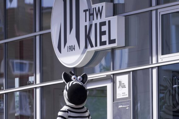 Eine Person in einem Zebrakostüm steht vor einem modernen Gebäude mit großen Fenstern und einem Schild mit der Aufschrift THW KIEL 1904.