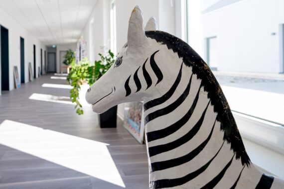 Eine bemalte Zebra-Skulptur steht in einem hellen Flur mit großen Fenstern, grauem Fußboden und Pflanzen an der Wand. Das Sonnenlicht fällt ein und wirft Schatten auf den Boden.