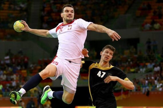 Ein Handballspieler in einem weiß-roten Trikot springt, um den Ball zu werfen, während ein Verteidiger in einem schwarz-gelben Trikot versucht, ihn während eines Spiels in einer großen Halle zu blockieren.