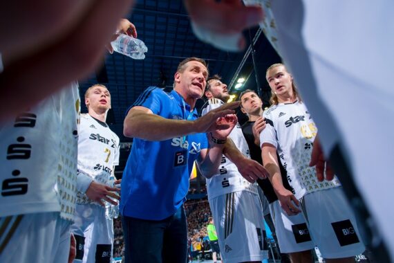Ein Trainer in einem blauen Hemd spricht während einer Auszeit leidenschaftlich zu einer Handballmannschaft in weißen Trikots, während die Spieler in einer Sportarena aufmerksam zuhören.
