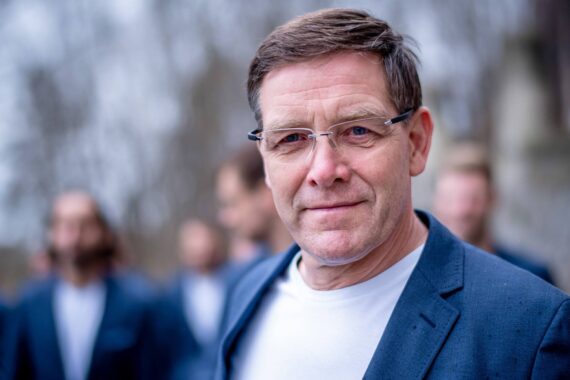 Ein Mann mittleren Alters mit kurzen braunen Haaren, Brille und einem blauen Blazer über einem weißen Hemd steht im Freien und lächelt leicht. Der Hintergrund ist unscharf und zeigt andere Menschen und Bäume.