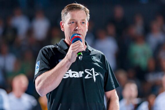 Ein Mann in einem schwarzen Polohemd mit weißen Logos spricht bei einer Indoor-Veranstaltung in ein Mikrofon mit der Aufschrift NDR, während das Publikum im Hintergrund verschwommen zu sehen ist.