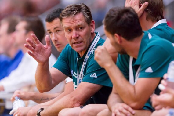 Ein Trainer in einem blaugrünen Hemd gestikuliert, während er bei einer Hallensportveranstaltung zu seiner Mannschaft auf der Bank spricht. Die Spieler und das Personal um ihn herum hören aufmerksam zu, mit Wasserflaschen in den Händen.