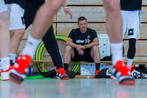 Ein Trainer sitzt auf einer Bank in einer Turnhalle und beobachtet, wie Sportler in Sportkleidung und mit leuchtend roten Schuhen an ihm vorbeiziehen. Hinter ihm stapeln sich Hula-Hoop-Reifen und eine Strategietafel an einer Holzverkleidung.