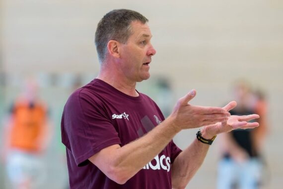 Ein Mann in einem kastanienbraunen Sporthemd gestikuliert mit seinen Händen, während er spricht, und scheint Anweisungen in einem Haus zu geben. Der Hintergrund ist unscharf, und es sind undeutliche Personen zu erkennen.