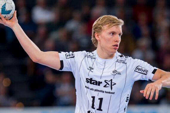 Ein Handballspieler, der ein weißes Trikot mit der Nummer 11 trägt, bereitet sich darauf vor, den Ball während eines Spiels zu werfen; im Hintergrund sind unscharfe Zuschauer zu sehen.