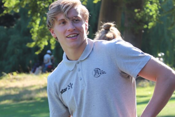 Ein junger Mann mit blonden Haaren und einem hellgrauen Poloshirt steht im Freien, lächelt und schaut zur Seite. Sonnenlicht und grüne Bäume bilden einen hellen, natürlichen Hintergrund.