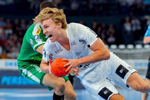 Ein männlicher Handballspieler in einem weißen Trikot stürzt sich mit einem orangefarbenen Ball in der Hand nach vorne, während ein Verteidiger in einem grünen Trikot versucht, ihn während eines intensiven Spiels zu blockieren.