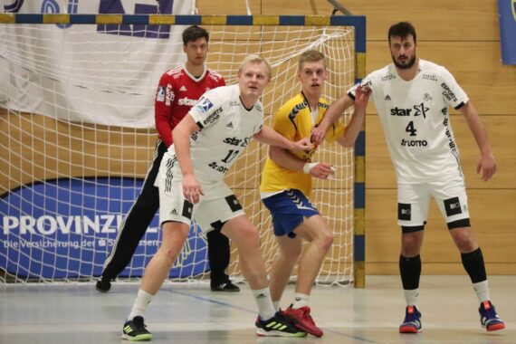 Drei Handballspieler in Weiß und einer in Gelb stehen vor einem Tor und bereiten sich auf das Spiel vor. Ein Torwart in Rot steht wachsam vor dem Netz und beobachtet das Geschehen genau.