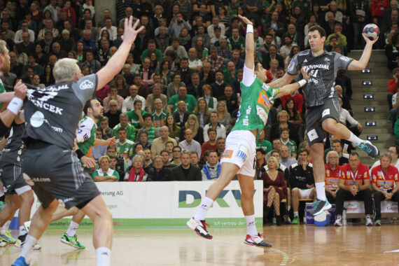 Ein Handballspieler in einem dunklen Trikot springt zum Schuss, während ein Spieler in einem grünen Trikot versucht, ihn abzublocken; die Zuschauer auf der Tribüne schauen gespannt zu.