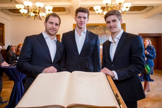 Drei junge Männer in Anzügen stehen lächelnd hinter einem offenen Gästebuch mit Unterschriften. Sie befinden sich in einem eleganten Raum mit Kronleuchtern, Holzböden und anderen Menschen im Hintergrund.