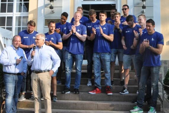 Eine Gruppe von Männern in blauen Hemden und Jeans steht auf einer Freitreppe und applaudiert, während zwei Männer, von denen einer ein Mikrofon hält, zu der Gruppe sprechen. Im Hintergrund ist ein Gebäude mit großen Fenstern und einer Tür zu sehen.