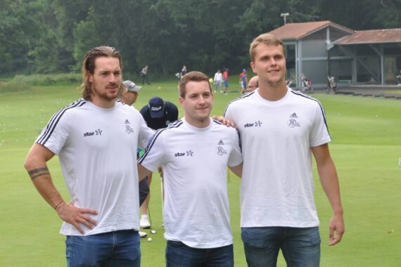 Drei Männer stehen zusammen auf einem Golfplatz und posieren für ein Foto. Sie tragen passende weiße T-Shirts mit Logos und Jeans. Im Hintergrund sind weitere Personen, eine Golfausrüstung und ein Gebäude zu sehen.