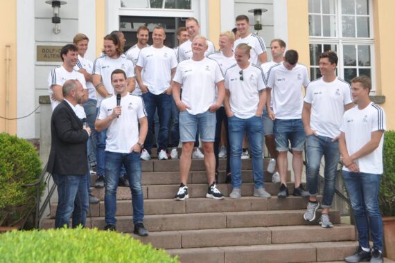 Eine Gruppe von Männern in passenden weißen Hemden und blauen Shorts oder Jeans steht lächelnd auf einer Freitreppe vor einem Gebäude, einige reden und lachen. Ein Mann hält ein Mikrofon in die Gruppe.