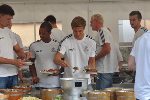 Eine Gruppe junger Männer in passenden weißen Sporthemden bedient sich an einem Buffettisch in einem weißen Zelt. Auf dem Tisch stehen verschiedene Gerichte und Salate in Metall- und Holzbehältern.
