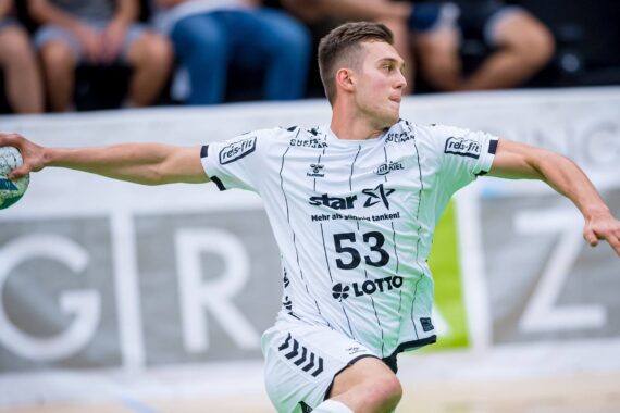 Ein männlicher Handballspieler in einem weiß-schwarzen Trikot mit der Nummer 53 bereitet sich darauf vor, den Ball während eines Spiels zu werfen. Im Hintergrund sind unscharfe Zuschauer und eine Fahne zu sehen.