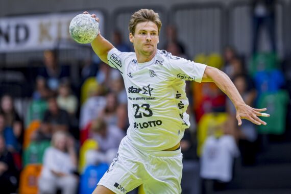 Ein männlicher Handballspieler in einem weißen Trikot bereitet sich darauf vor, den Ball während eines Spiels zu werfen. Er ist mitten in der Aktion, mit erhobenem Arm und konzentriertem Blick, während die Zuschauer von der Tribüne im Hintergrund zusehen.