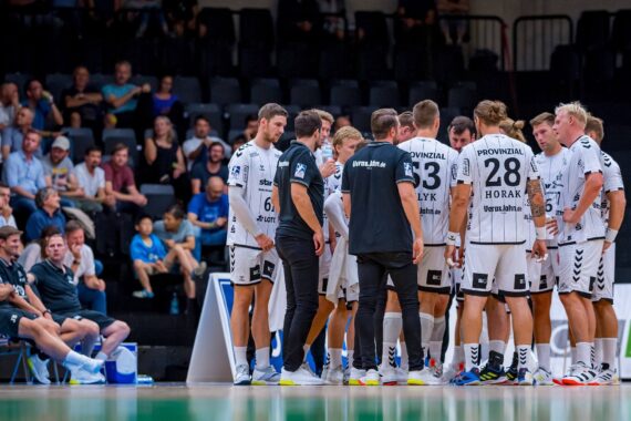Eine Herren-Handballmannschaft in weißen Trikots steht während einer Auszeit auf einem Hallenplatz mit den Trainern zusammen, während die Zuschauer im Hintergrund von der Tribüne aus zusehen.