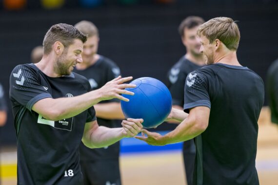 Zwei Männer in schwarzer Sportkleidung lächeln und halten gemeinsam einen blauen Medizinball in einer Turnhalle, mit anderen Personen im Hintergrund, was auf ein Teamtraining hindeutet.