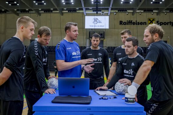 Ein Trainer in einem blauen Hemd bespricht mit sechs männlichen Handballspielern in schwarzen Trikots, die in einer Sporthalle um einen blauen Tisch mit Laptop, Wasser und Handbällen versammelt sind, eine Strategie.