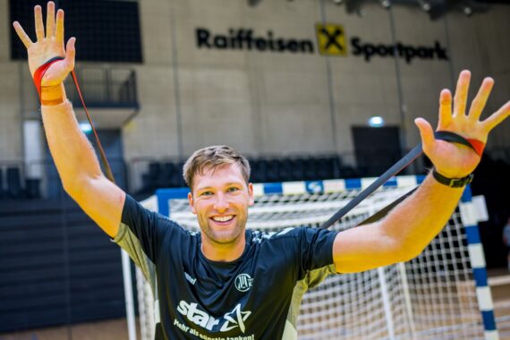 Ein lächelnder Mann in Sportkleidung dehnt ein Widerstandsband mit beiden Händen vor einem Handballtor in einer Halle im Raiffeisen Sportpark.