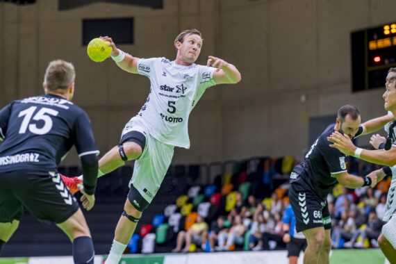 Ein Handballspieler in einem weißen Trikot springt, um den Ball zu werfen, während drei Gegner in schwarzen Trikots ihn verteidigen. Das Spiel findet in einer Halle mit leeren bunten Sitzen im Hintergrund statt.
