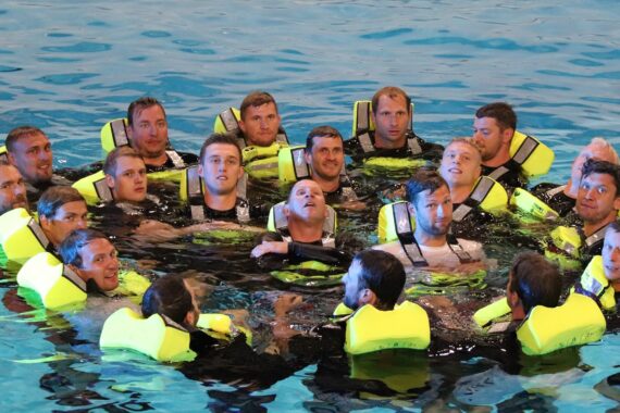 Eine Gruppe von Männern, die gelbe Schwimmwesten tragen, bilden einen engen Kreis, während sie gemeinsam in einem Schwimmbecken schwimmen, nach oben schauen und während einer Wasser-Sicherheits- oder Überlebenstrainingsübung aufmerksam wirken.