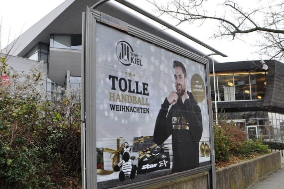 Auf einer Plakatwand vor einem modernen Gebäude ist eine Weihnachtswerbung für den THW Kiel Handball zu sehen, die einen lächelnden Mann in einem Mannschafts-Sweatshirt, ein Plüsch-Maskottchen und den Text TOLLE HANDBALL WEIHNACHTEN zeigt.