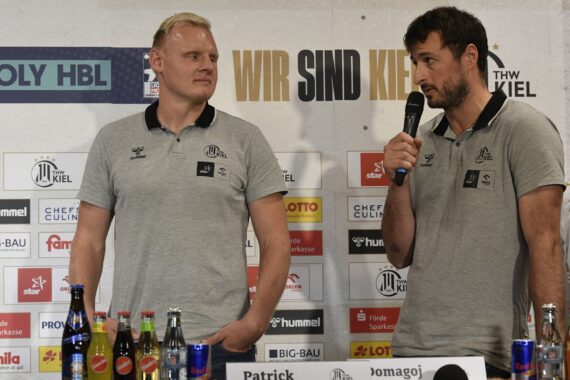Zwei Männer in grauen Poloshirts stehen sich bei einer Pressekonferenz mit Mikrofonen gegenüber. Auf dem Tisch vor ihnen stehen verschiedene Getränke. Im Hintergrund sind Logos und der Schriftzug WIR SIND KIEL zu sehen.