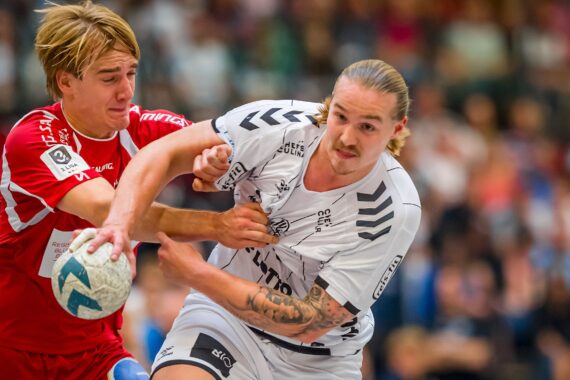 Zwei männliche Handballspieler kämpfen intensiv um den Ball; der eine im roten Trikot versucht, den Ball des anderen im weißen Trikot zu blockieren oder zu stehlen, während eines Hallenspiels mit einer unscharfen Menschenmenge im Hintergrund.