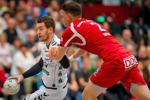 Zwei männliche Handballspieler in Aktion: Der eine im weißen Trikot hält den Ball und bewegt sich vorwärts, während der andere in Rot versucht, ihn zu verteidigen. Eine unscharfe Menge schaut im Hintergrund zu.