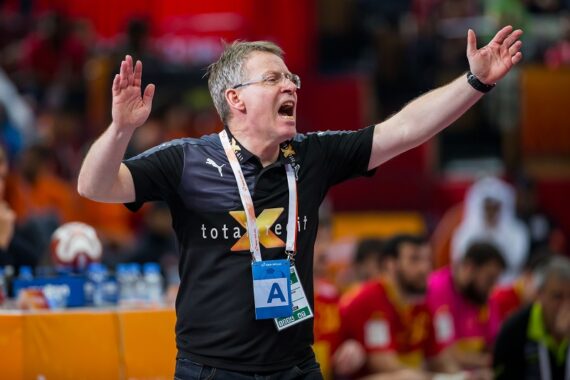 Ein Trainer, der eine Brille und ein schwarzes Hemd trägt, steht mit erhobenen Armen da und scheint während eines Handballspiels Anweisungen zu rufen, während im Hintergrund Spieler und Zuschauer zu sehen sind.