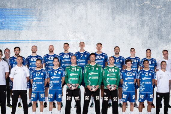 Eine Herren-Handballmannschaft in blauen Trikots steht in zwei Reihen mit Trainern und Betreuern in weißen Hemden vor einem grauen Hintergrund mit dem Logo des VfL Gummersbach auf der linken Seite.