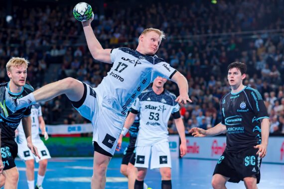 Ein Handballspieler in einem weiß-grauen Trikot springt während eines Spiels zum Wurf, umringt von gegnerischen Spielern in schwarzen und blauen Trikots, während im Hintergrund eine Menschenmenge zuschaut.