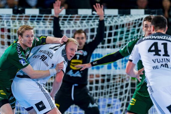 Ein Handballspieler in einem weißen Trikot versucht, sich in der Nähe des Tores von zwei Verteidigern in grünen Trikots zu befreien, während ein Torwart mit erhobenen Armen vor dem Netz steht.