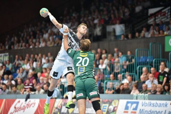 Ein Handballspieler in einem weißen Trikot springt zum Wurf, während ein Verteidiger im grünen Trikot mit der Nummer 20 versucht, ihn zu blockieren. Die Aktion findet in einer überfüllten Halle statt, in der Zuschauer zuschauen.