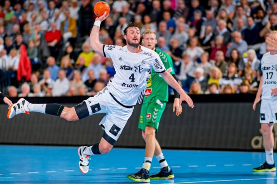 Ein Handballspieler in einem weißen Trikot springt mit einem orangefarbenen Ball durch die Luft und versucht zu werfen, während ein Verteidiger in Grün zuschaut; eine Zuschauermenge füllt den Hintergrund.