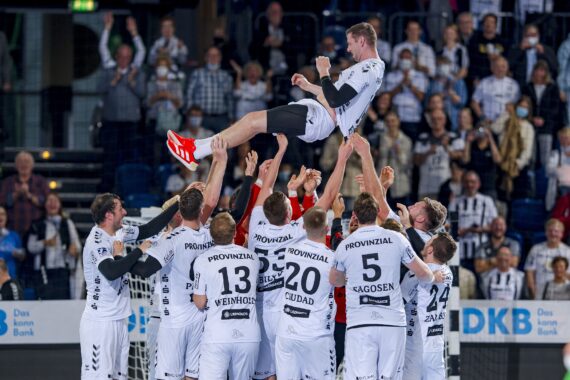 Eine Gruppe von Handballern in weißen Trikots hebt einen Mannschaftskameraden in die Luft, umringt von jubelnden Fans auf der Tribüne.
