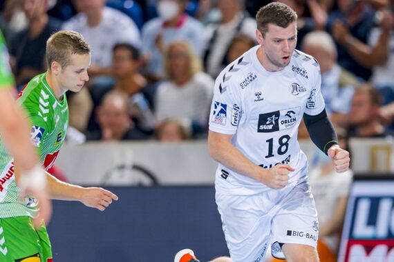 Zwei männliche Handballspieler in Aktion während eines Spiels. Der Spieler in Weiß, der die Trikotnummer 18 trägt, scheint zu jubeln, während der Spieler in Grün nach unten schaut. Im Hintergrund ist eine unscharfe Zuschauermenge zu sehen.