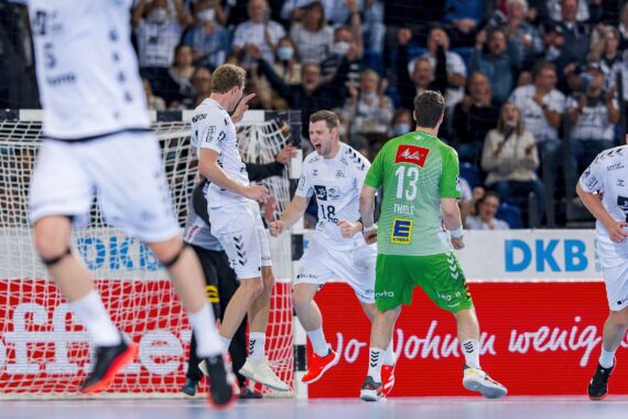 Handballspieler in weißen Trikots feiern ein Tor auf dem Spielfeld, während ein Spieler in einem grünen Trikot neben dem Tor steht. Eine jubelnde Menge füllt die Tribüne im Hintergrund.