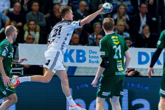 Ein Handballspieler in einem weißen Trikot springt und bereitet sich auf den Wurf vor, während zwei Verteidiger in grünen Trikots zusehen. Das Spiel findet in einer Halle mit Zuschauern im Hintergrund statt.