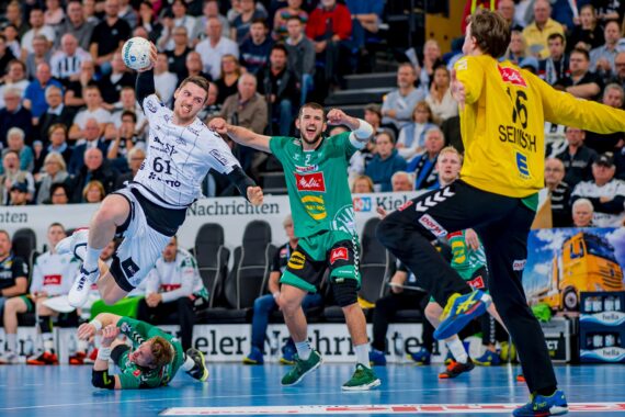 Ein Handballspieler in Weiß springt zum Schuss, während er von Spielern in Grün verteidigt wird; der Torwart in Gelb versucht, den Schuss zu parieren. Die Zuschauer im Hintergrund schauen gebannt zu.
