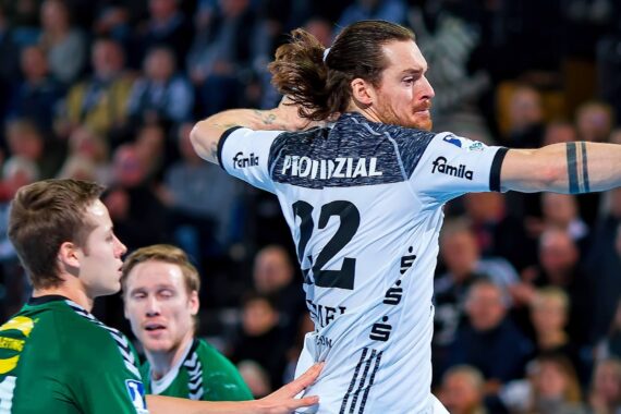 Ein männlicher Handballspieler in einem weißen Trikot ist im Begriff, den Ball zu werfen, während zwei gegnerische Spieler in grünen Trikots versuchen, ihn während eines Spiels in einer überfüllten Halle zu blockieren.